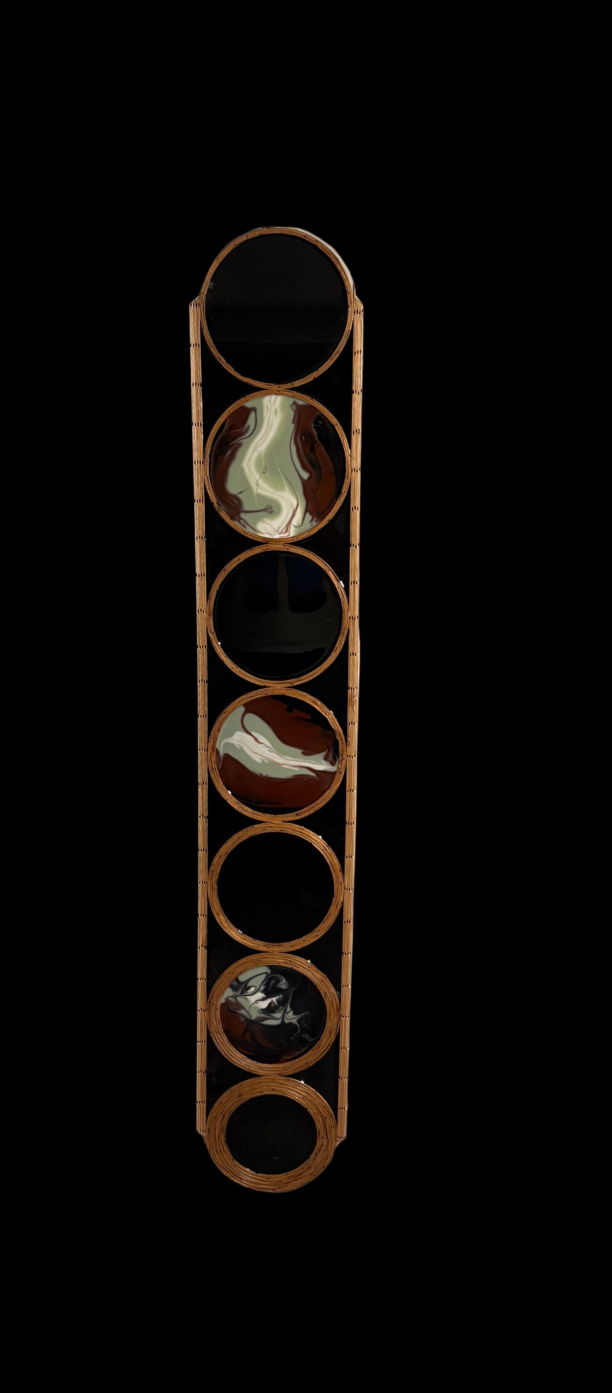 Tall vertical sculpture of seven stacked circles made of craft sticks. Three hold swirled resin panels in green, white and rust; four are left as black voids.